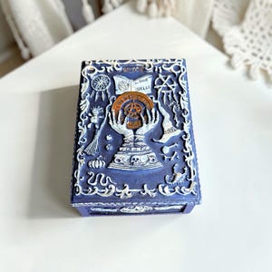 Könnte beinhalten: Dunkelblaue, rechteckige Box mit erhabenem, weißem, verziertem Design. Die Box zeigt ein zentrales Design einer Kristallkugel, die von Händen gehalten wird, mit den Worten "Witch Book of Spells" darüber. Weitere Elemente sind ein Besen, ein Hut und ein Kürbis.