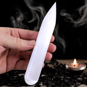 May include: A white selenite wand with a pointed tip, held in a hand against a dark background with smoke and candles.
