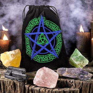 May include: A black velvet pouch with a green and blue pentagram embroidered on the front. The pouch is surrounded by various crystals, including a yellow citrine, a green aventurine, a pink rose quartz, and a black obsidian.