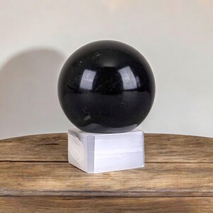 Puede incluir: Una esfera de obsidiana negra sobre un soporte blanco. La esfera es lisa y pulida. El soporte est&aacute; hecho de un material blanco.