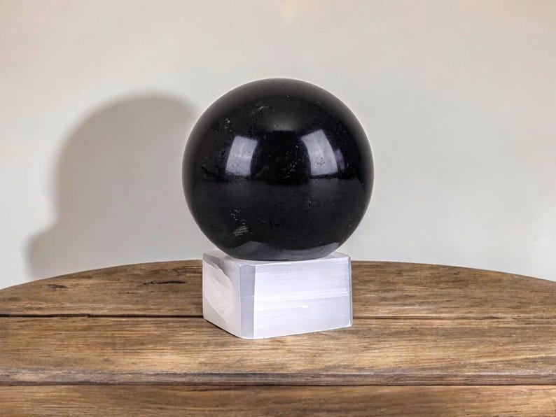 Puede incluir: Una esfera de obsidiana negra sobre un soporte blanco. La esfera es lisa y pulida. El soporte est&aacute; hecho de un material blanco.
