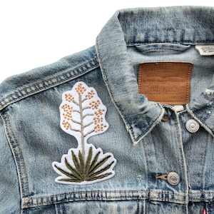 May include: A blue denim jacket with a white embroidered patch of a yucca plant with brown flowers. The jacket has a brown leather label that says "Levi Strauss & Co. Genuine Quality Clothing" and a size tag.