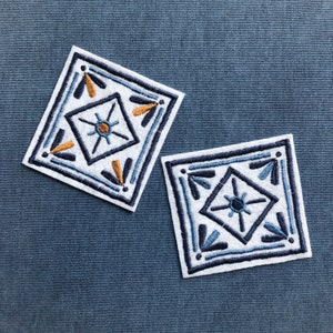 May include: Two white square fabric patches with blue and orange embroidery. The embroidery features a geometric pattern with a star in the center.
