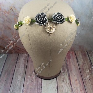 May include: A floral crown with black and white roses, cream-colored flowers, and green leaves. The crown is displayed on a wooden surface.