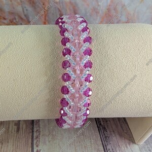 May include: A beaded bracelet featuring alternating rows of pink and magenta beads, with smaller clear beads in between. The bracelet is displayed on a neutral-colored surface, showcasing its intricate design and color contrast. The bracelet is a piece of jewelry.