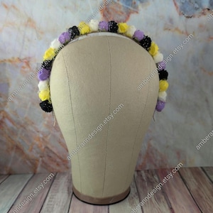 May include: A floral headband featuring small, colorful flowers in yellow, white, purple, and black. The flowers are arranged around a thin, silver-colored band. The headband is displayed on a mannequin head.