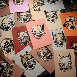 Könnte beinhalten: Eine Sammlung farbenfroher Kunstkarten mit skurrilen, monsterartigen Gesichtern. Jede Karte hat ein einzigartiges Design mit schwarzen Umrissen und gemalten Details in Weiß, Beige und Braun. Die Karten sind auf einer dunklen Oberfläche angeordnet.