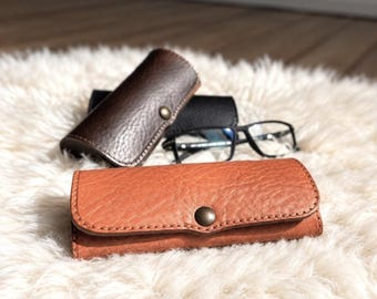 May include: Three rectangular cases with snap closures and a pair of black-framed spectacles. One case is light tan, one is dark brown, and one is black. Each case features a textured surface and a single metal snap button on the front flap.