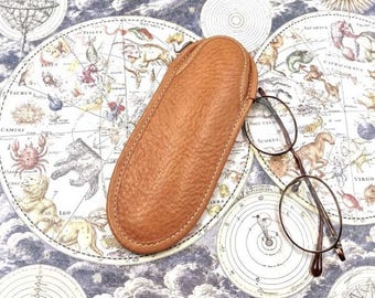 May include: A tan, textured, oval-shaped leather eyeglass case with visible stitching along the edges, positioned next to a pair of thin-rimmed, dark brown metal glasses on a background featuring a vintage celestial star map.
