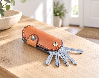 May include: A tan leather key organizer featuring a fold-over design with visible stitching along the edges. The case is secured by a silver-toned metal snap button and a brass-colored screw post, holding a set of five silver metal keys.