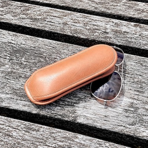 Può includere: Un astuccio per occhiali in pelle color cuoio con un esterno liscio e arrotondato e cuciture visibili lungo i bordi, posizionato accanto a un paio di occhiali da sole aviatore con montatura argentata e lenti scure che riflettono il cielo.