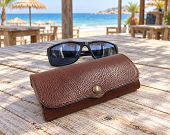May include: A dark brown leather glasses case with a pebbled texture and a single brass-coloured metal snap button closure, positioned in front of a pair of black-framed sunglasses on a wooden surface.