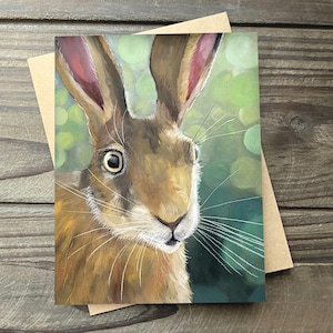 May include: A greeting card featuring a detailed painting of a hare's face. The hare has brown fur, large eyes, and long whiskers. The background is a soft green with bokeh effect. The card is paired with a brown envelope.