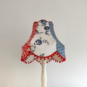 May include: A white table lamp with a red, white and blue patterned lampshade. The lampshade has a scalloped edge and a red pom-pom trim.