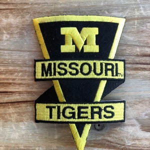 May include: Embroidered patch featuring the Missouri Tigers logo. The patch is black with yellow accents, including the letter "M" and the words "MISSOURI TIGERS". The patch is in a shield shape with a pointed bottom.