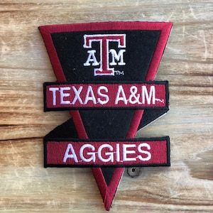 May include: A black and maroon triangular patch with the letters "T" "A" "M" in white, the text "TEXAS A&M" in white on a maroon background, and the word "AGGIES" in white on a maroon background.