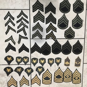 May include: A collection of military rank insignia patches in various shades of green and gold. The patches feature different chevron designs and an eagle emblem.