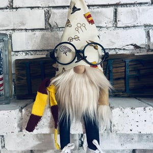 May include: A whimsical gnome doll with a cream-colored cone hat adorned with Harry Potter-themed illustrations. The gnome wears round black glasses, a long white beard, and a scarf in burgundy and gold. It has blue pants and white sneakers.