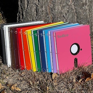Könnte beinhalten: Ein Stapel von 15 bunten Notizbüchern mit schwarzen Umschlägen und silbernen Ringen. Die Notizbücher sind in einem Regenbogenmuster angeordnet, wobei das letzte Notizbuch rosa ist und das Wort "Sudico" sowie ein schwarz-weißes Grafikdesign aufweist.