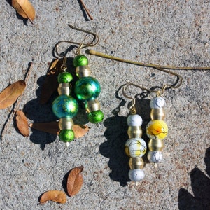 May include: Two pairs of dangle earrings with gold-toned metal and glass beads. One pair has green and blue glass beads, and the other pair has white and yellow glass beads.