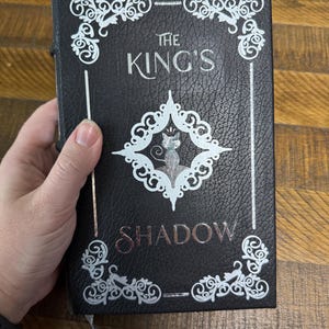 Puede incluir: Libro negro titulado "The King's Shadow" con letras y adornos plateados. La portada presenta una ilustraci&oacute;n de gato plateado dentro de un dise&ntilde;o en forma de diamante. Se adjunta un marcador de cinta plateada.