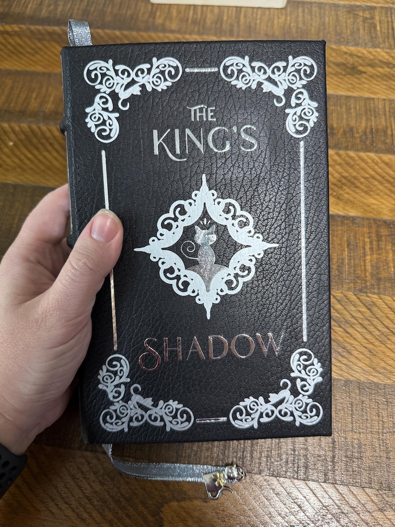 Puede incluir: Libro negro titulado "The King's Shadow" con letras y adornos plateados. La portada presenta una ilustraci&oacute;n de gato plateado dentro de un dise&ntilde;o en forma de diamante. Se adjunta un marcador de cinta plateada.