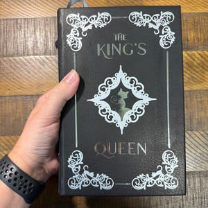 Puede incluir: Libro negro con el t&iacute;tulo "THE KING'S QUEEN" en letras plateadas y cobrizas. La portada presenta adornos blancos y una silueta de gato. Una cinta azul de marcador es visible en la parte superior.
