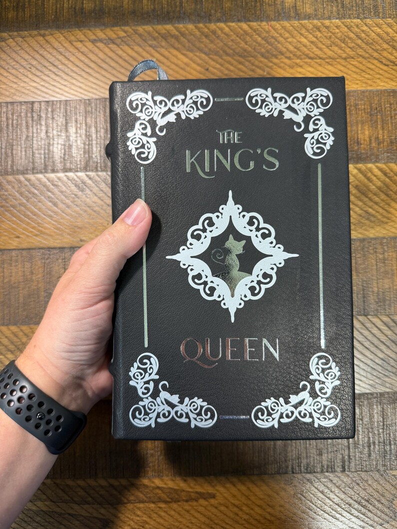 Puede incluir: Libro negro con el t&iacute;tulo "THE KING'S QUEEN" en letras plateadas y cobrizas. La portada presenta adornos blancos y una silueta de gato. Una cinta azul de marcador es visible en la parte superior.