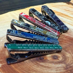 Könnte beinhalten: Sechs Metall-Krawattenklammern mit Leiterplatten-Designs in verschiedenen Farben. Die Klammern weisen rote, blaue, grüne und schwarze Designs auf. Die Krawattenklammern sind auf einer Holzoberfläche angeordnet.