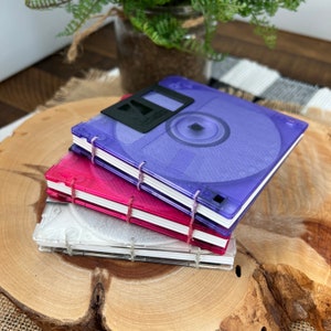 Peut inclure: Trois carnets empilables fabriqués à partir de disquettes réutilisées. Le carnet du haut est violet, le carnet du milieu est rose et le carnet du bas est blanc. Le carnet du haut porte le texte "Verbatim" sur la face avant.