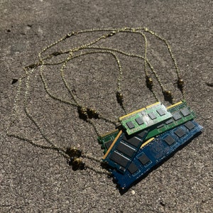 May include: Three green and blue computer memory sticks are tied together with a gold chain necklace. The necklace has multiple strands and is decorated with small gold beads.