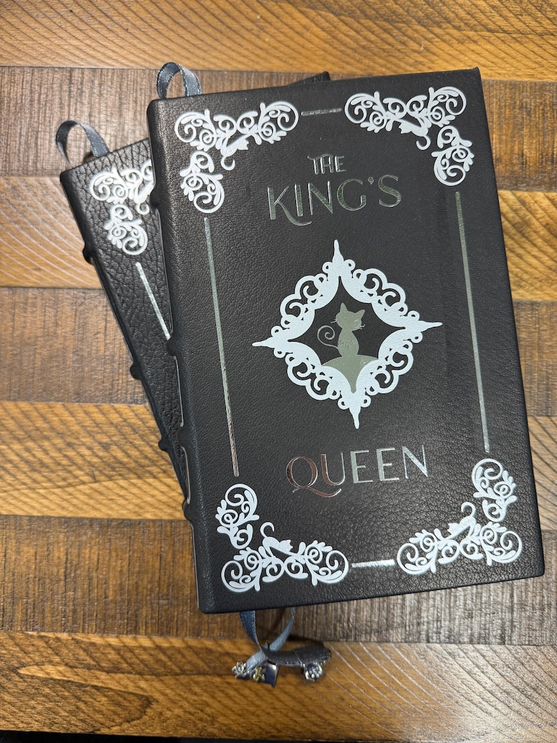 Puede incluir: Dos diarios negros con el texto plateado "THE KING'S QUEEN". Presentan esquinas decorativas de filigrana blanca y un dise&ntilde;o central con una silueta de gato. Cada diario tiene un marcap&aacute;ginas de cinta azul.