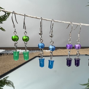 Puede incluir: Cinco pares de pendientes con ganchos plateados y cuentas de vidrio de colores. Las cuentas son verdes, azules y moradas.