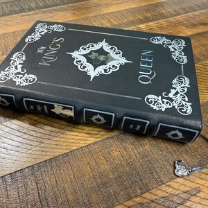 Puede incluir: Un libro negro con el texto "The King's Queen" en letras plateadas y filigranas blancas decorativas. Una marca de libro plateada con un peque&ntilde;o dije est&aacute; adjunta. El libro est&aacute; sobre una superficie de madera.