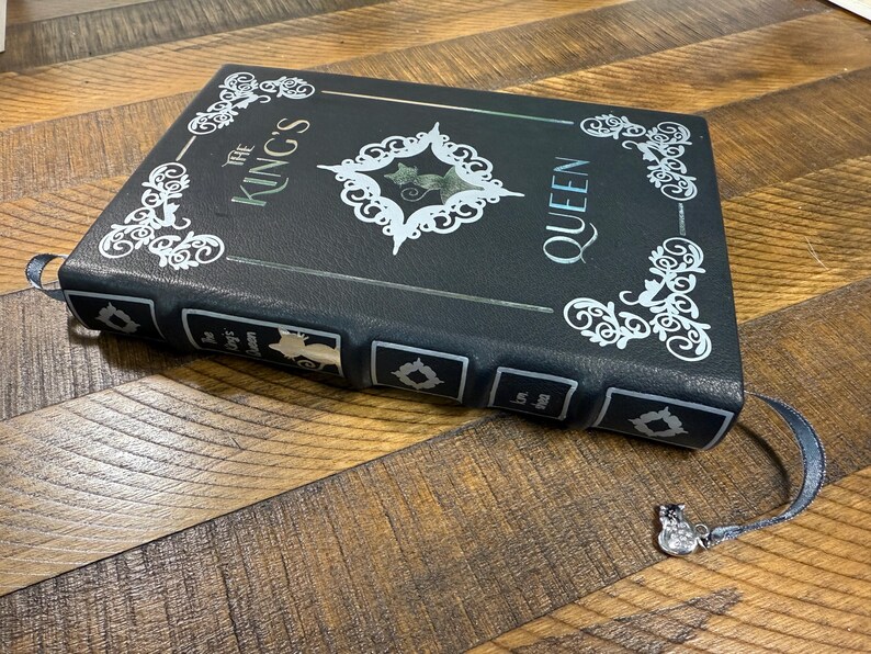 Puede incluir: Un libro negro con el texto "The King's Queen" en letras plateadas y filigranas blancas decorativas. Una marca de libro plateada con un peque&ntilde;o dije est&aacute; adjunta. El libro est&aacute; sobre una superficie de madera.