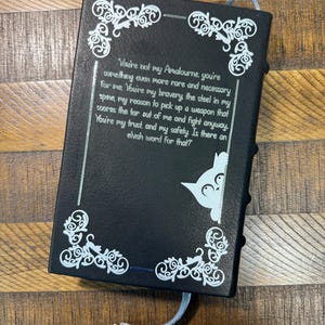 Puede incluir: Diario negro con dise&ntilde;os decorativos blancos en las esquinas y texto. Un gr&aacute;fico de gato azul claro asoma por el lado derecho. El diario tiene un marcap&aacute;ginas de cinta azul claro con un dije plateado.