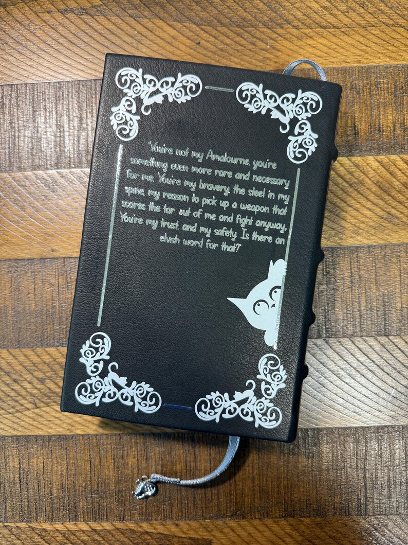 Puede incluir: Diario negro con dise&ntilde;os decorativos blancos en las esquinas y texto. Un gr&aacute;fico de gato azul claro asoma por el lado derecho. El diario tiene un marcap&aacute;ginas de cinta azul claro con un dije plateado.