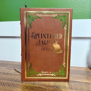May include: A brown book with gold lettering and accents. The title "Splintered Magic Series" is displayed with a cat silhouette. The book is bound with a bookmark and features the text "Books 1-3".