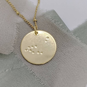 May include: A gold necklace with a round pendant featuring a pattern of raised dots. The pendant is hanging on a light gray fabric background.