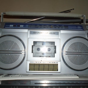 May include: Silver Sharp GF-4646 boombox with large speakers, a cassette player, and a radio dial. The boombox has a handle for carrying and an extendable antenna. The model name is visible on the front.