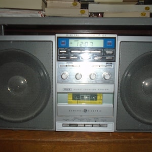 Op de afbeelding: Een vintage General Electric boombox met grote luidsprekers aan weerszijden van een centraal bedieningspaneel. Het digitale display toont de tijd als 12:07. De boombox is zilver en grijs, met een cassettespeler.