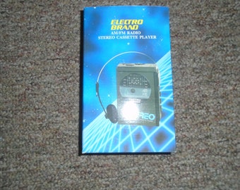Op de afbeelding: Een blauwe doos met een witte rand en een zwart-zilveren AM/FM radiocassetterecorder met koptelefoon. Op de doos staat "Electro Brand AM/FM Radio Stereo Cassette Player".