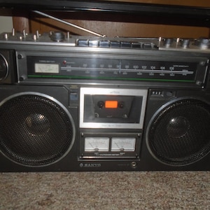 May include: A Sanyo portable stereo boombox featuring a black and silver chassis, two large circular speakers, a central cassette deck, a radio tuner with a frequency scale, and two analogue power meters located below the cassette compartment.
