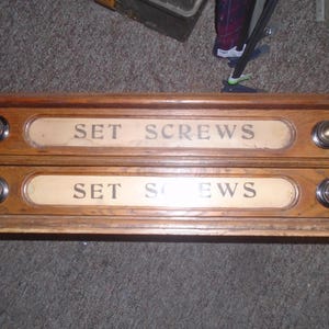 Può includere: Un mobiletto rettangolare in legno con due cassetti orizzontali. Ogni cassetto presenta un'etichetta color crema con la scritta SET SCREWS in nero e due pomelli rotondi in metallo color ottone sui lati.
