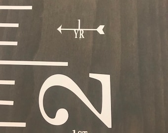 May include: A white wooden growth chart with black markings. The chart has a white arrow with the text "1 YR" and another white arrow with the text "1ST DAY" and the number "2" printed on it.