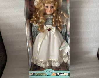 May include: A porcelain doll with blonde curly hair wearing a white dress with lace trim and a green floral print top. The doll is holding a small white book and is wearing a straw hat. The doll is in a box with the words "Noble Heritage Collection" and "A Special Edition" printed on it.