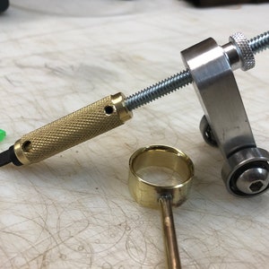 May include: A metal and brass jewelry-making tool set. The set includes a textured brass handle with a black tip, a silver screw mechanism, a brass ring, and a green plastic cap. The tools are on a light-colored surface.