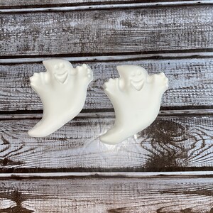 May include: Two white ghost-shaped decorations with smiling faces. The ghosts have a curved, flowing design, with outstretched arms. The background is a rustic wooden surface with horizontal planks, creating a contrast with the smooth, white ghosts. Halloween decor.