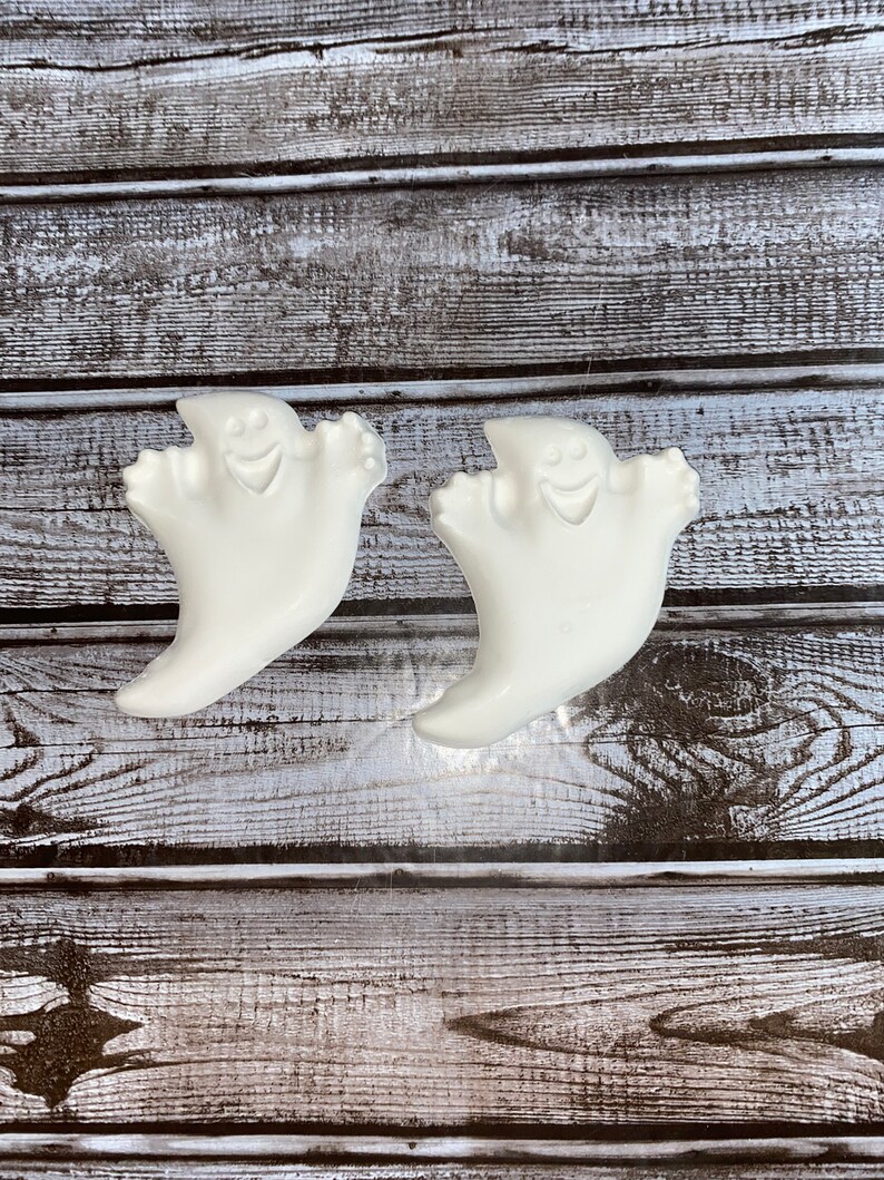 May include: Two white ghost-shaped decorations with smiling faces. The ghosts have a curved, flowing design, with outstretched arms. The background is a rustic wooden surface with horizontal planks, creating a contrast with the smooth, white ghosts. Halloween decor.