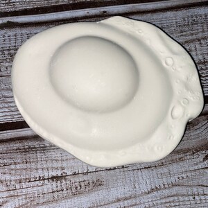 May include: A white ceramic dish shaped like a fried egg. The dish has a rounded center and a wavy edge with small, circular indentations. The egg-shaped dish is on a wooden surface.